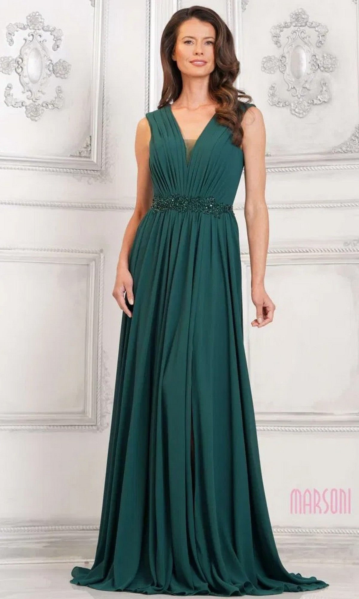 A woman in a Marsoni MV1292 long formal dress in deep green, featuring a sleeveless V-neck, ruched bodice, and beaded waistband, stands in an ornately decorated white room.
